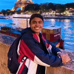 A person in a red, white and blue jacket wears a backpack and poses in front of the Eiffel Tower.