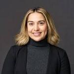 Alejandra wears a dark gray turtleneck and black blazer in front of a gray background. 