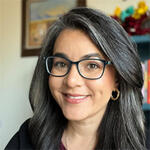 Vanessa smiling wearing glasses and sitting in front of framed paintings.